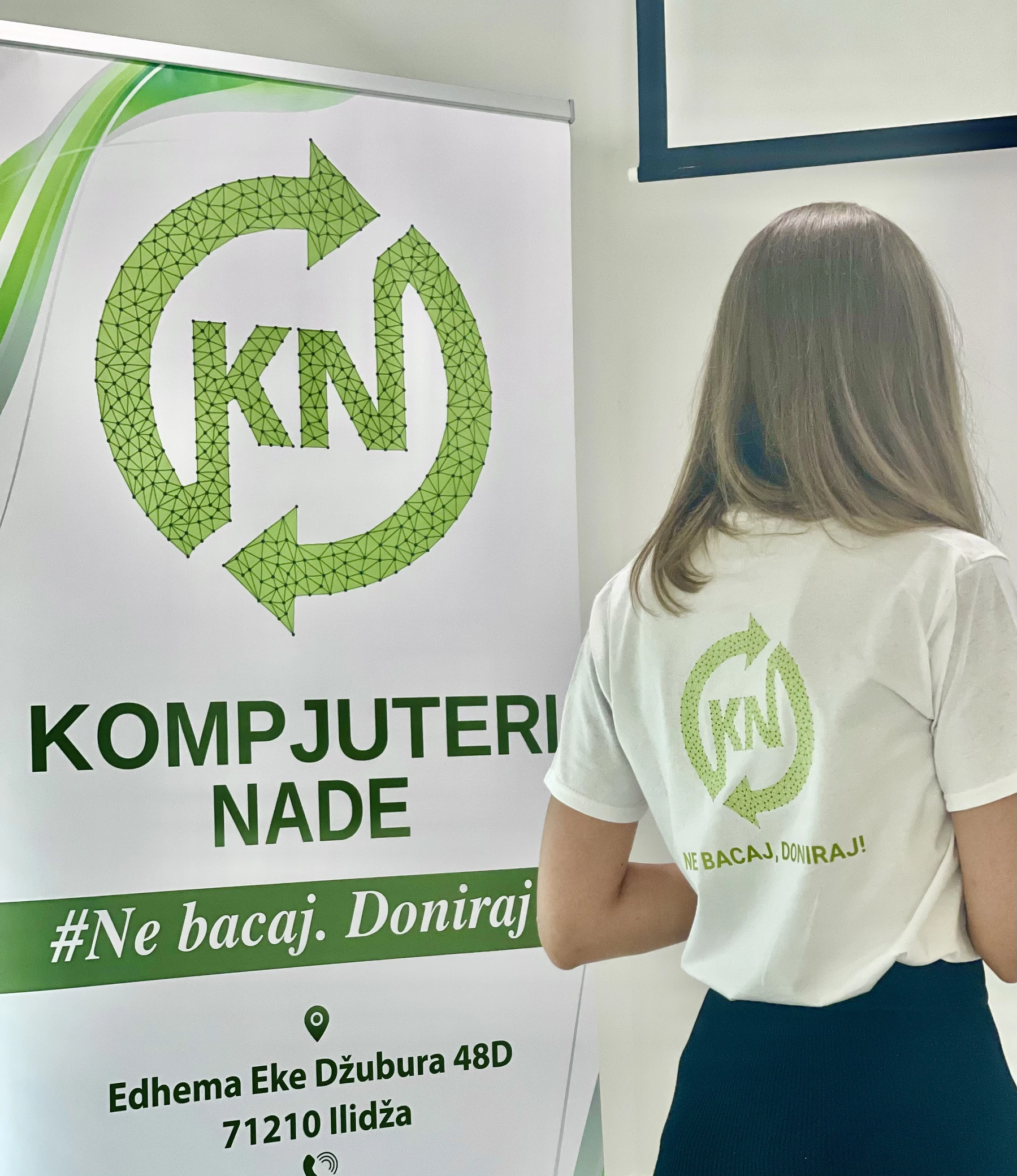 A volunteer wearing a Kompjuteri nade shirt beside the KN banner at a community event.